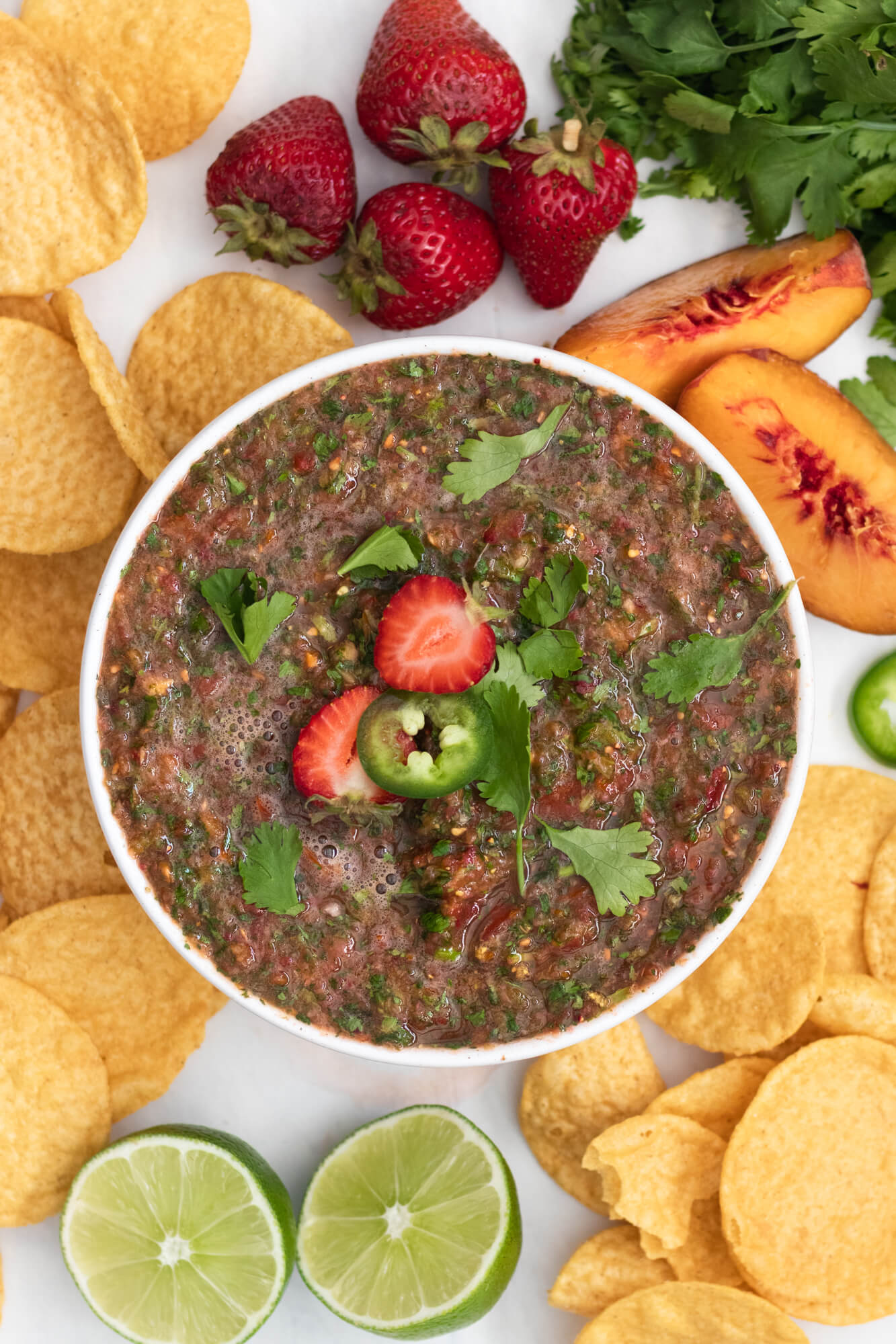 A bowl of strawberry peach salsa surrounded my chips and a halved peach, strawberries, cilantro and a sliced lime