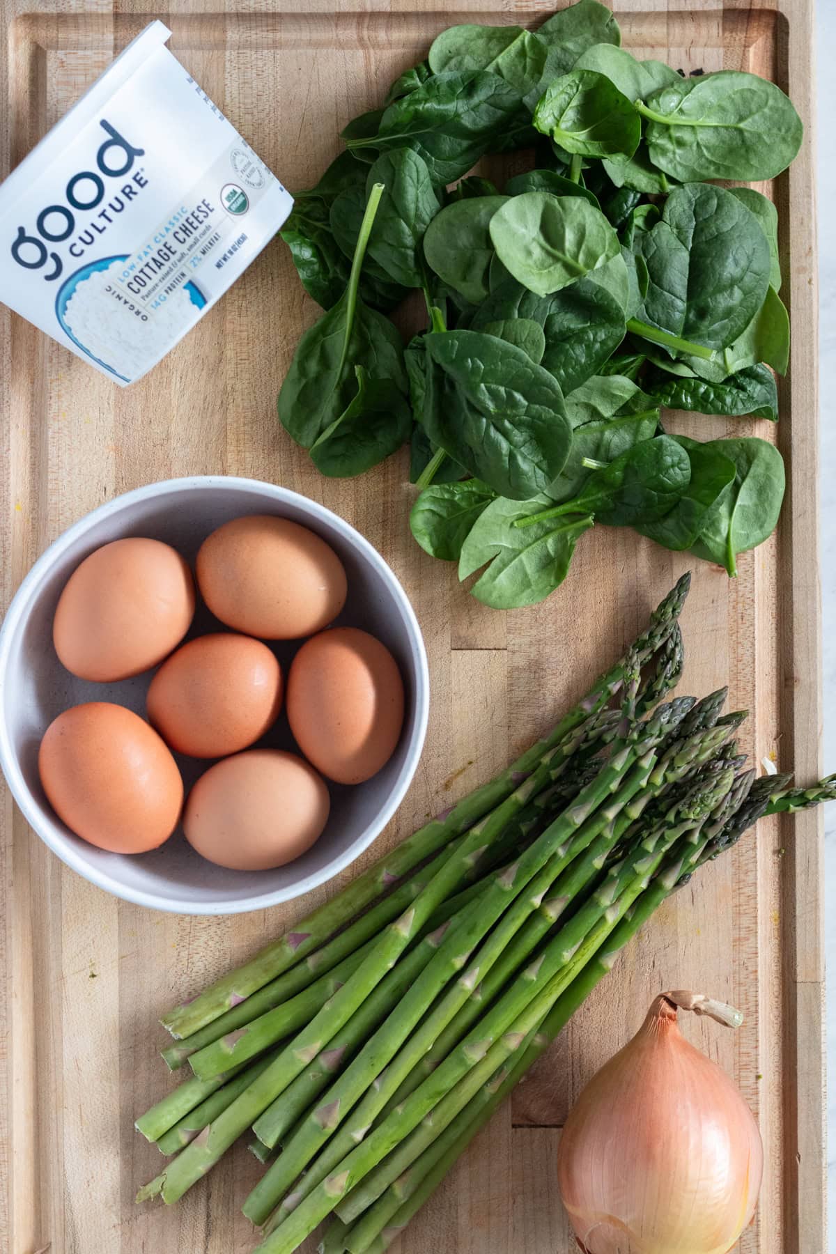 Eggs in a bowl with spears of asparagus, spinach, an onion and a container of cottage cheese.
