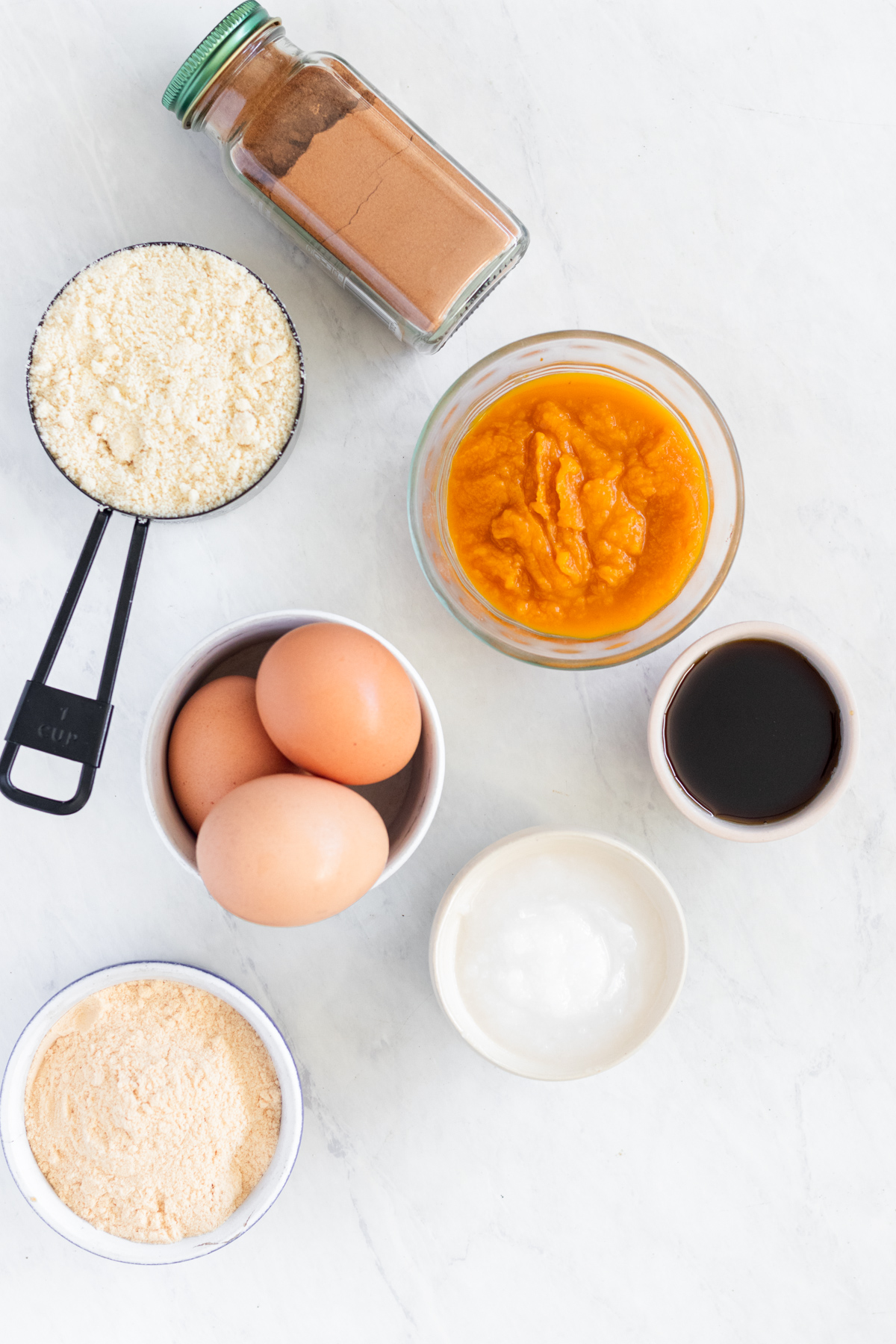 Small bowls with eggs, pumpkin puree, vanilla, baking powder and almond flour near pumpkin spice.