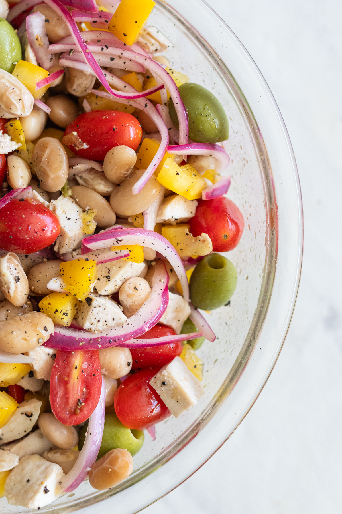 Up close, overhead view of Mediterranean Butter Bean Salad in a clear glass bowl, filled with cherry tomatoes, olives, diced bell pepper, and butter beans tossed in olive oil.